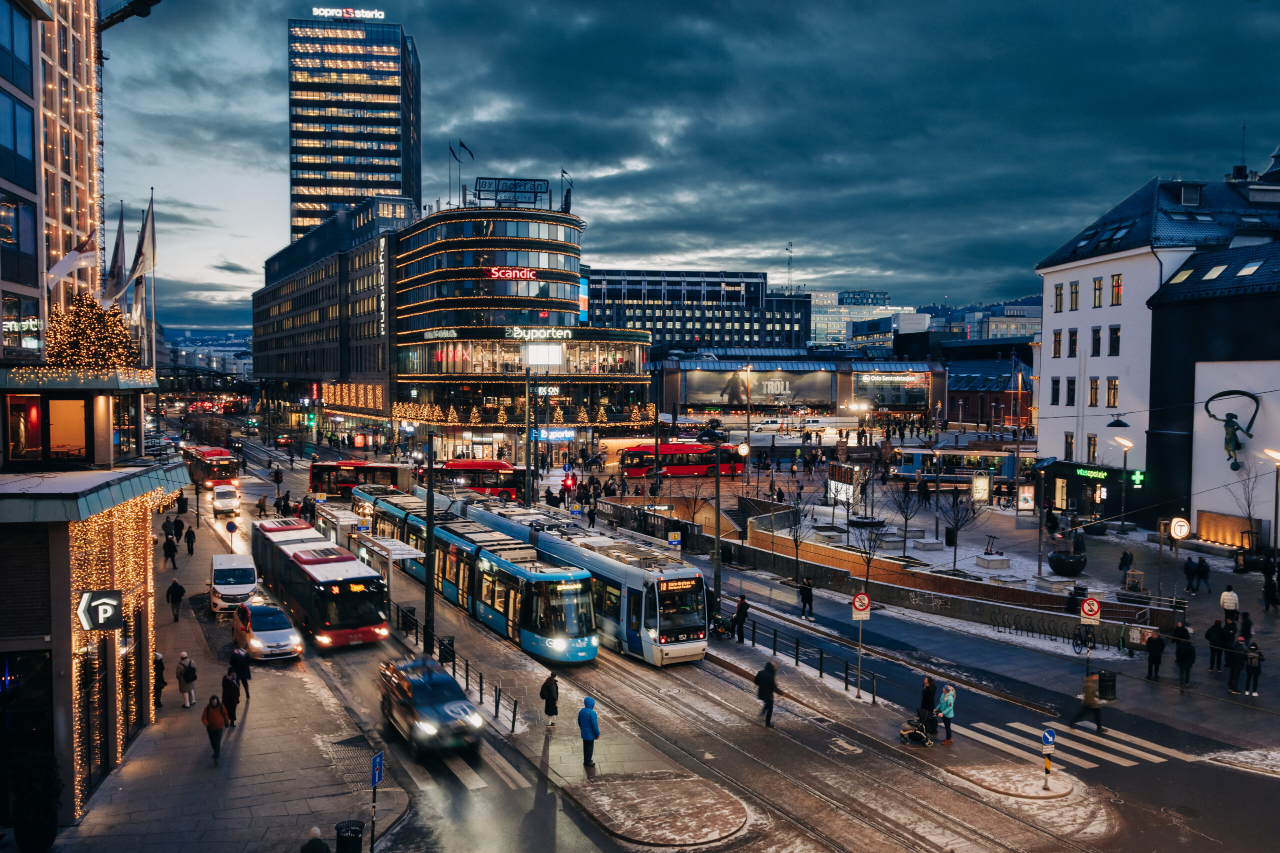 Biler, trikker og busser på Jernbanetorget omringet av bygg i skumringstid.