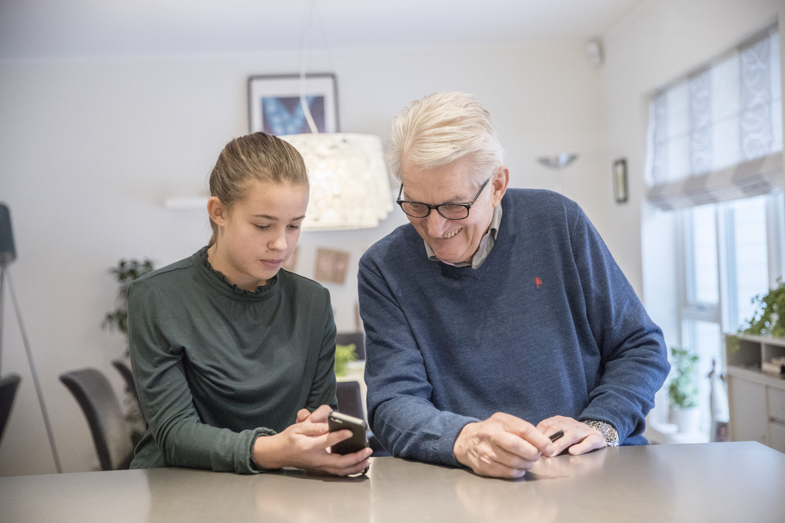 En ung jente viser en eldre mann skjermen på sin mobiltelefon, sittende på kjøkkenet.