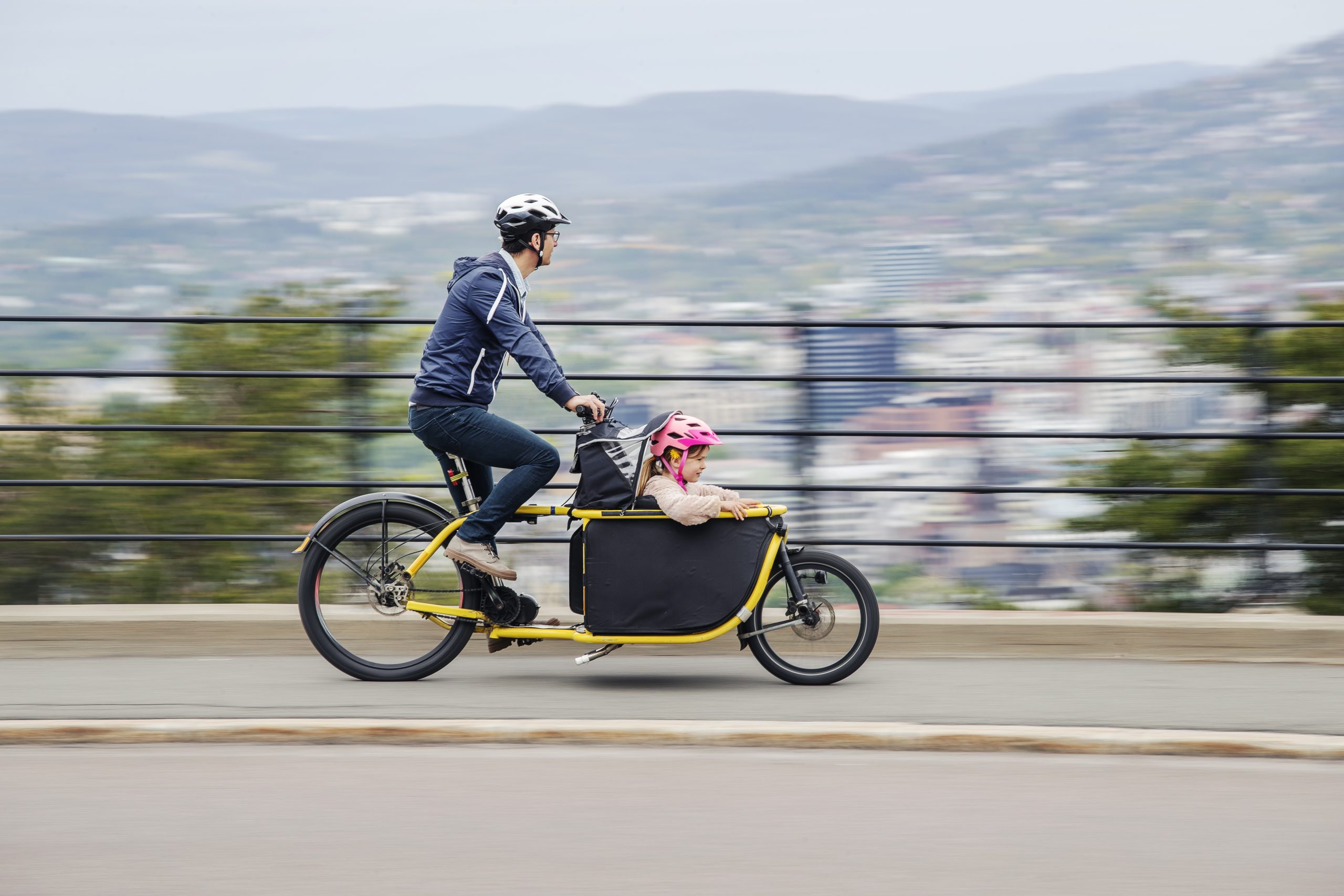 Mann med et barn sittende i et sete foran på sykkelen kjører nedover en bakke med Oslo som kan skimtes i bakgrunnen.