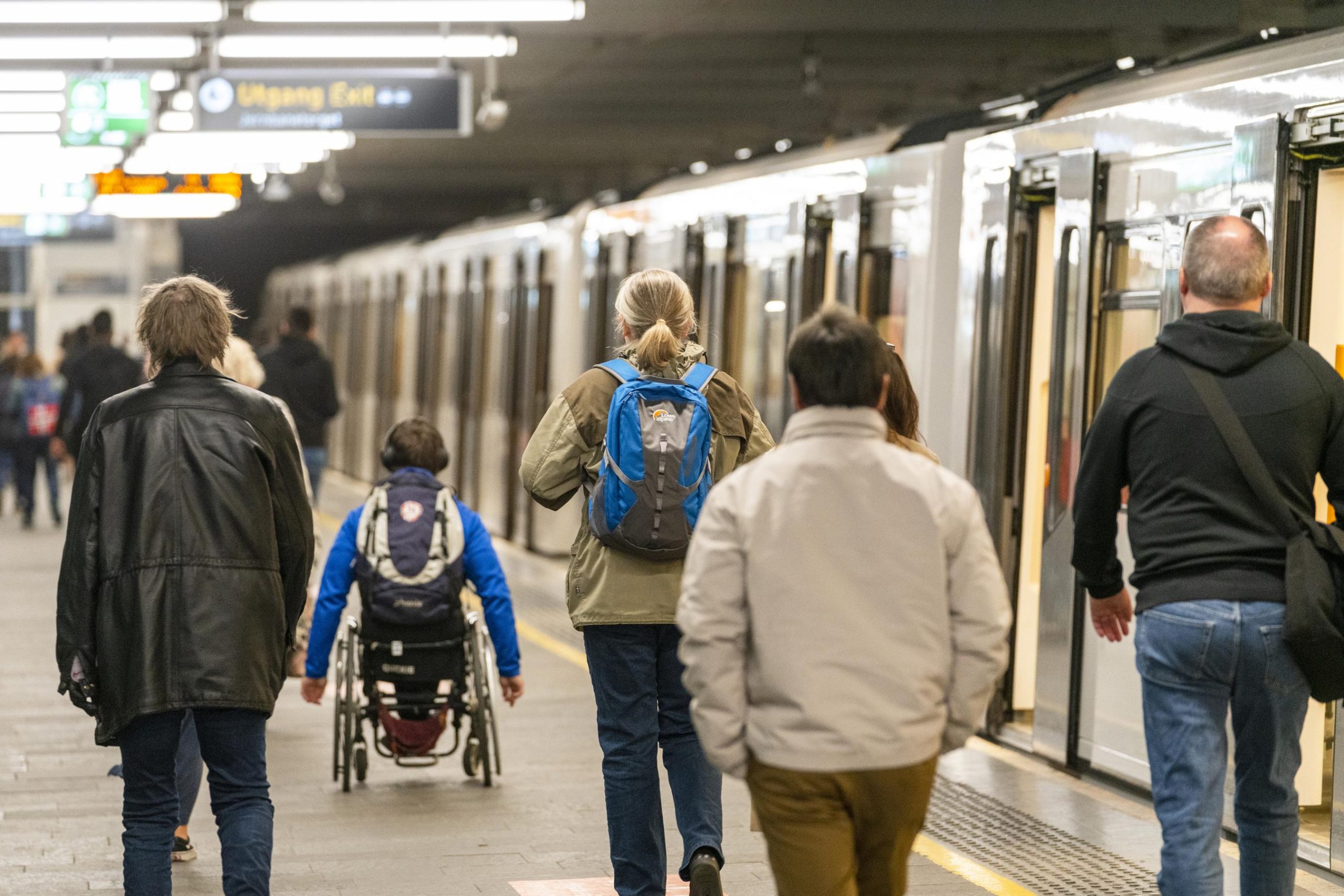 En person i rullestol og flere gående personer passerer en t-bane inne på en t-banestasjon.