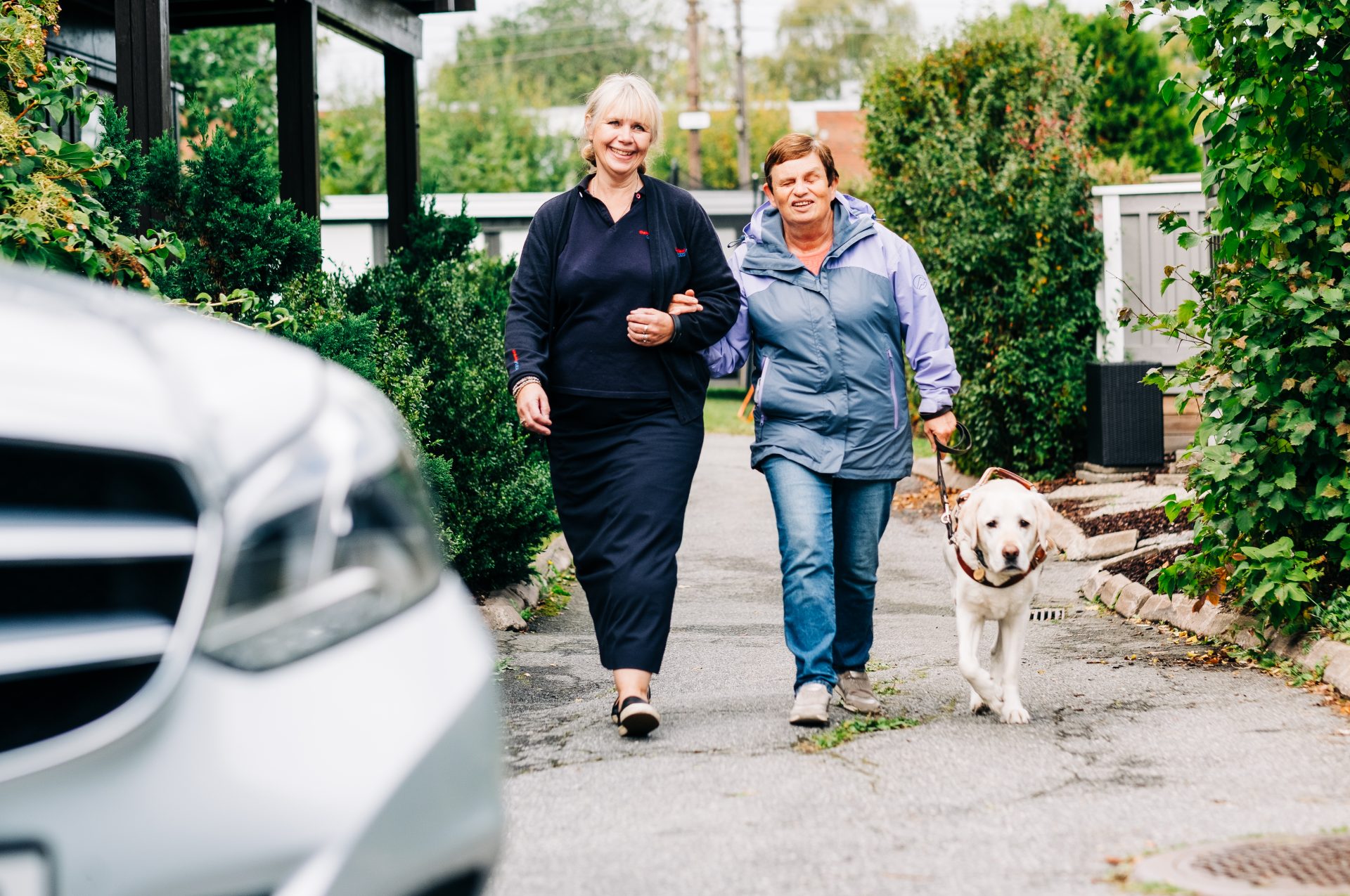 Blind kvinne med førerhund blir ledsaget fra huset sitt, og mot en bil, av en annen kvinne