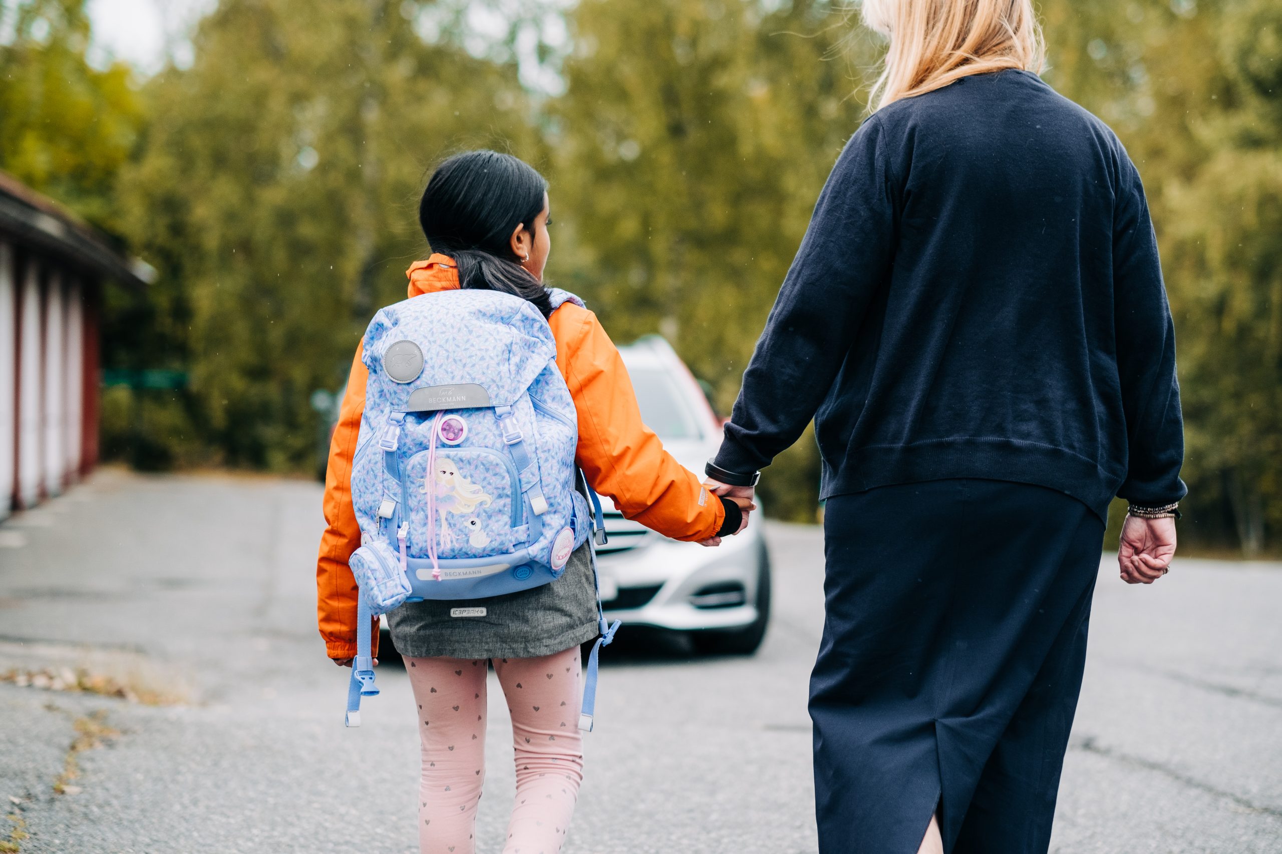 Mor, og barn med skolesekk leier mens de går bortover en vei.