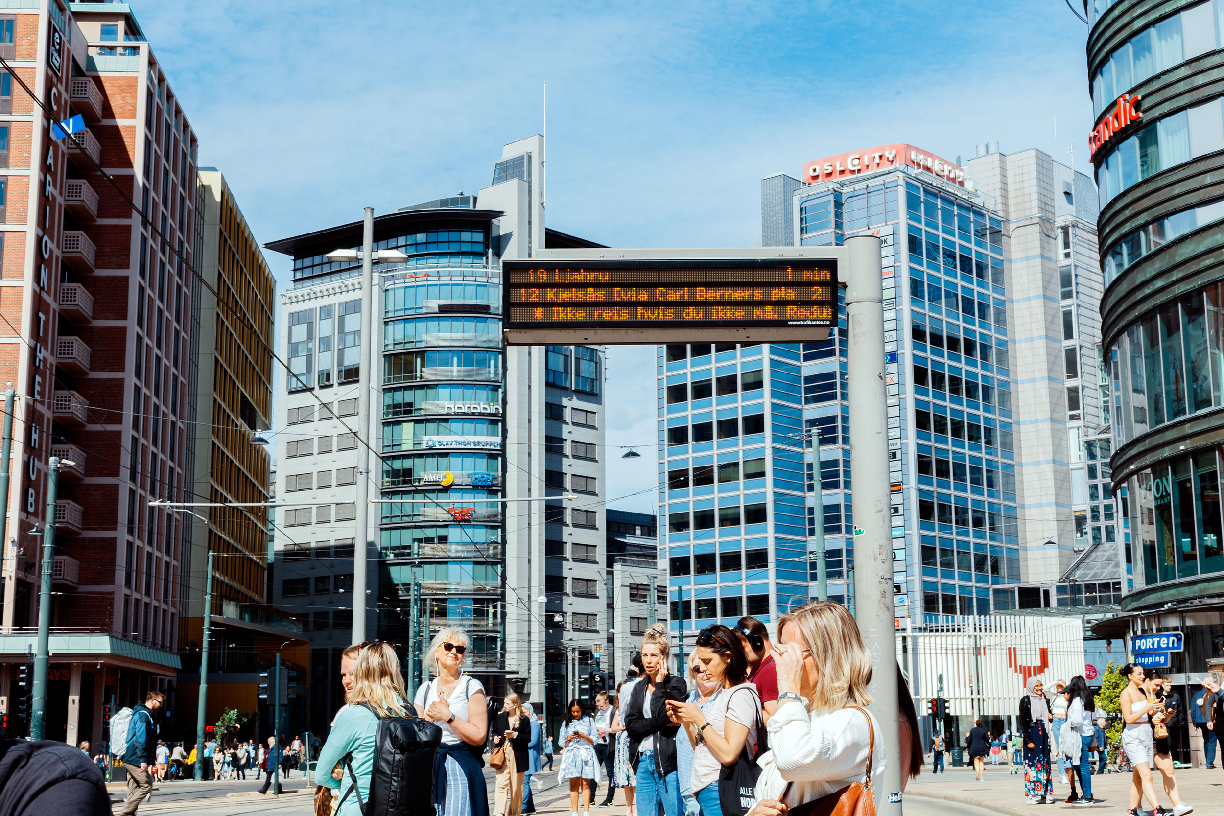 Reisende som venter på kollektivtransport på Jernbanetorget. Informasjon om smittervern står på sanntidsskilt.