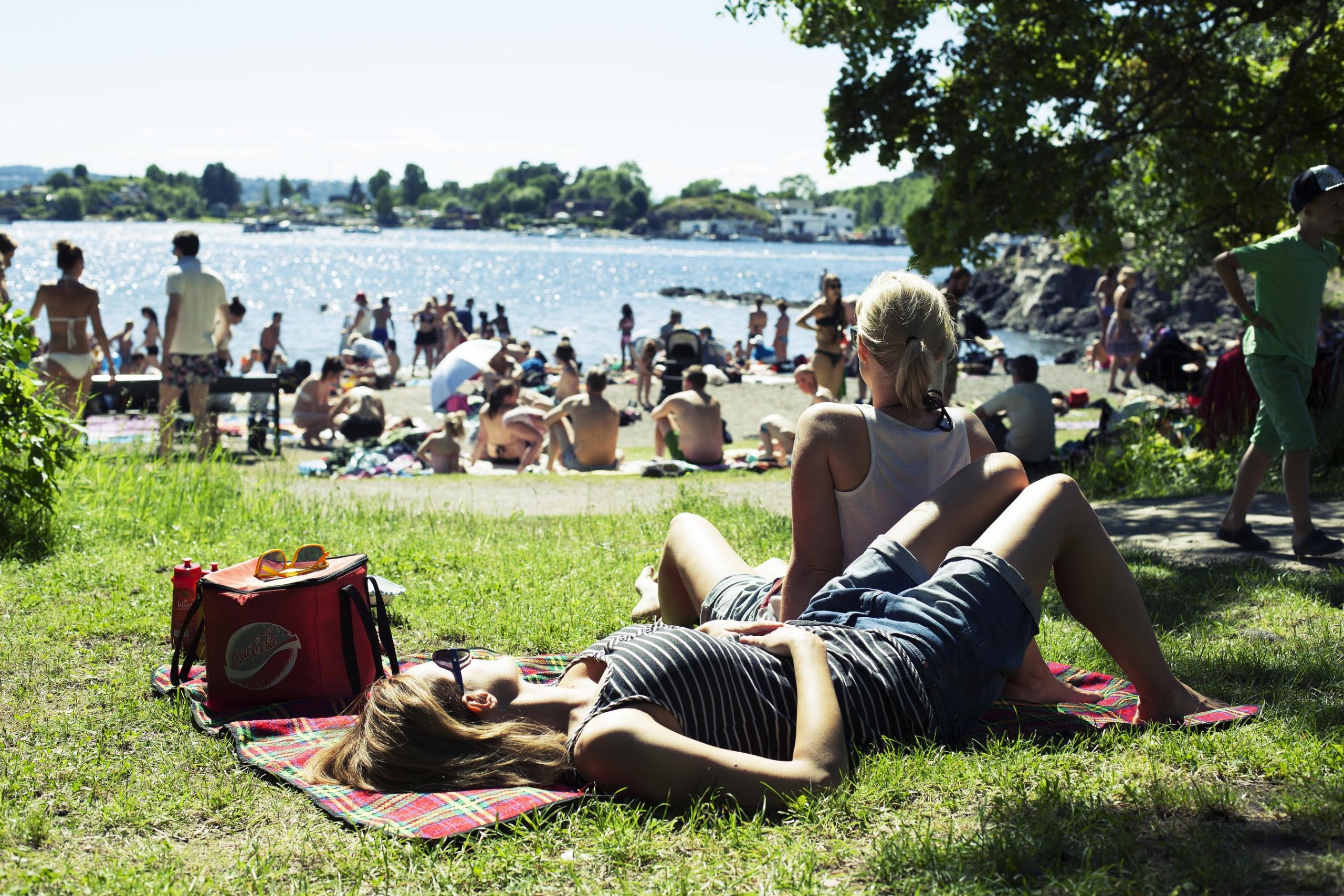 Two women sunbathe on the grass and look out over a crowded beach on a summer day.