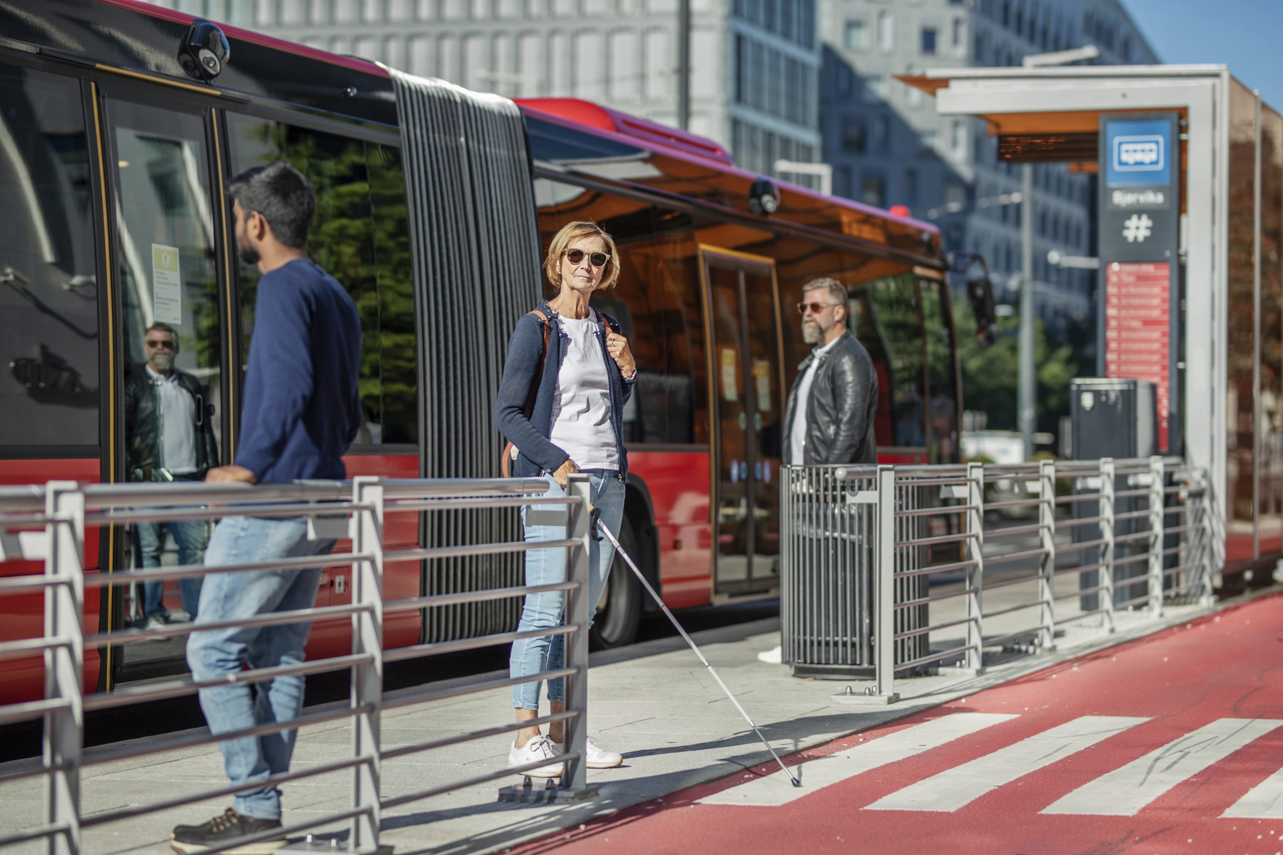 Blind woman with a cane at a bus stop in an urban environment with a bus in the background.