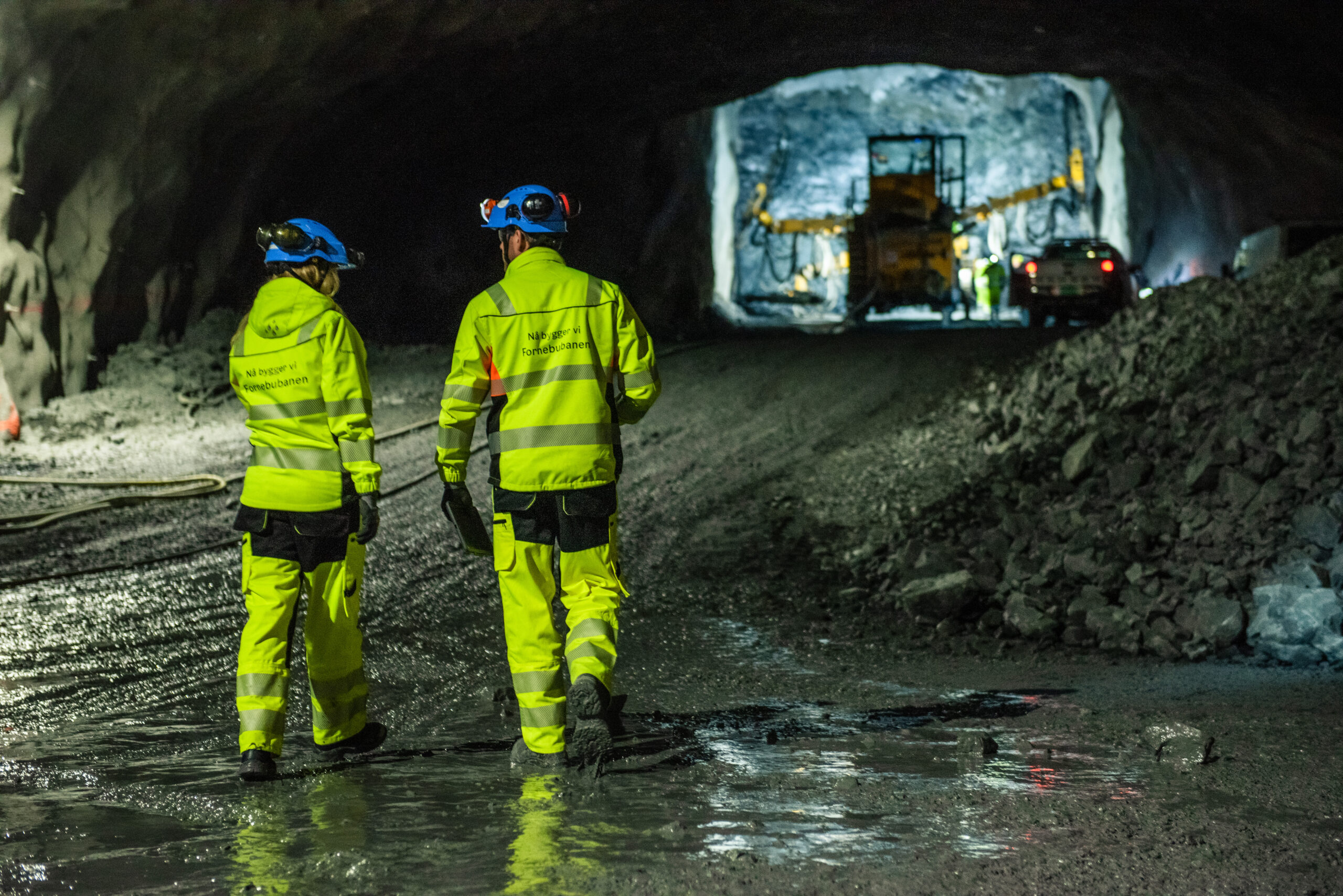 Two men in yellow and blue protective gear are talking in a tunnel. They are placed in the foreground of a truck and a car.