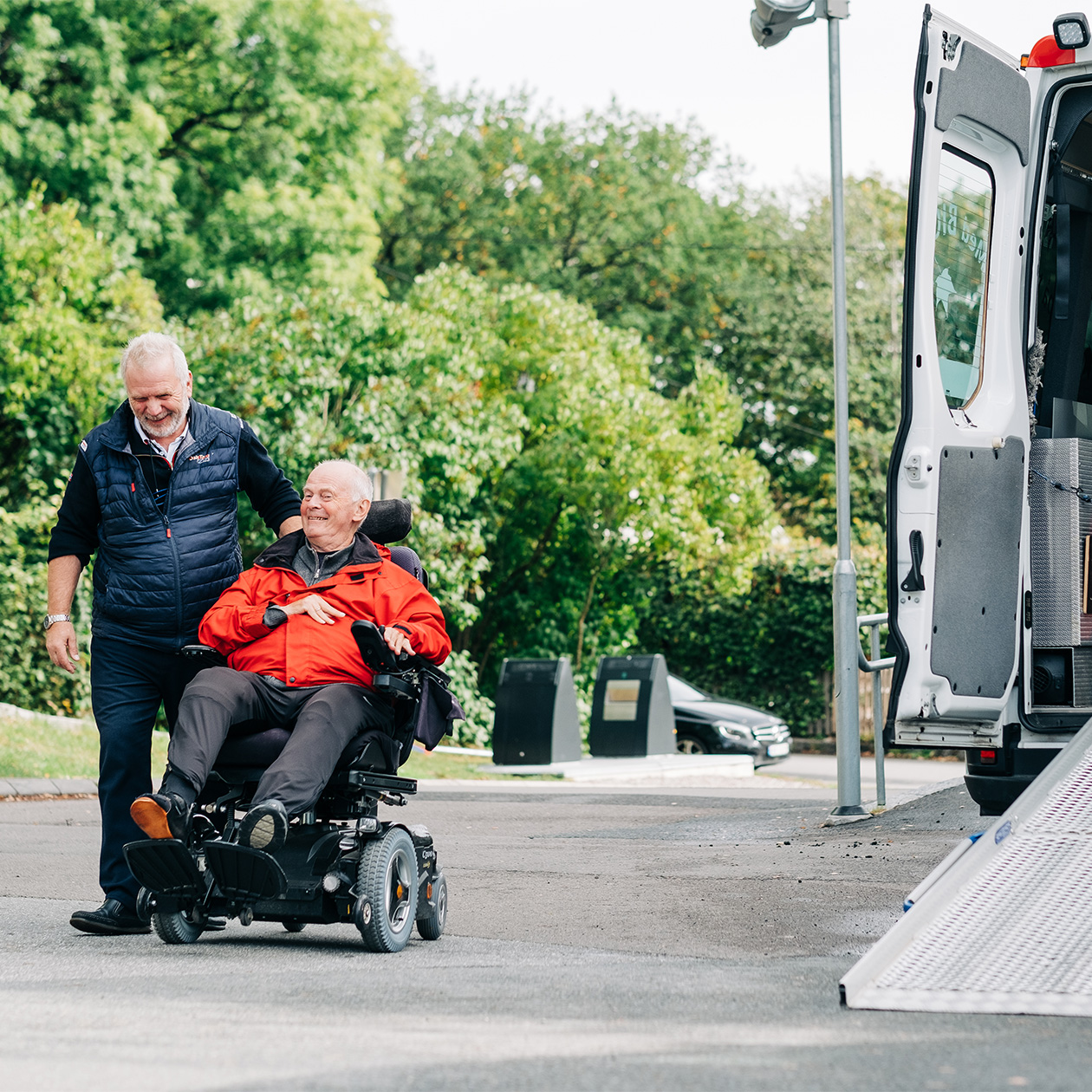 Man in an electric wheelchair is helped to the vehicle ramp by the driver.